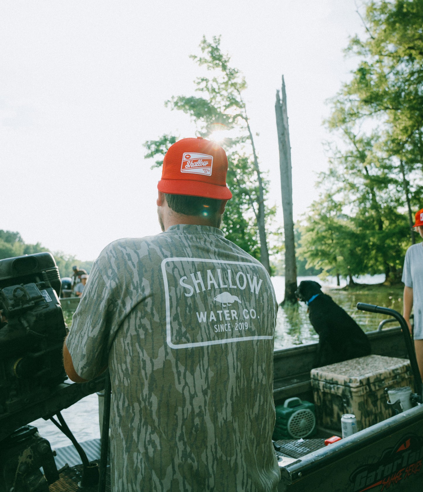 Person wearing a Mossy Oak Camo Shallow Water Co. Logo T-shirt and red All mesh cap on a boat, with trees and water in the background.