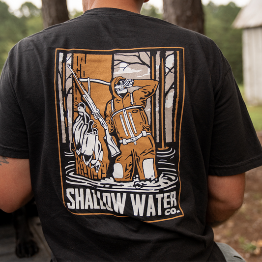 Person wearing the Killing Time Tee by Shallow Water Co. in a wooded, swampy environment—design showing a skeleton hunter in waders with ducks over the shoulder, embodying the spirit of waterfowl hunting.